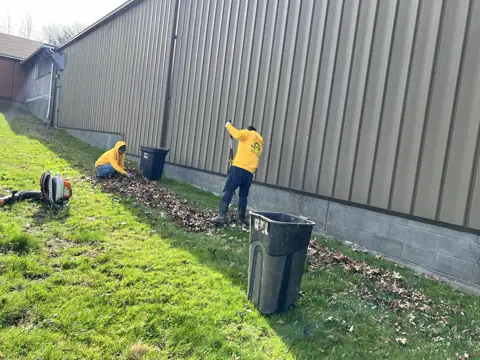 A crew member cleaning leaves from a bed beside a building
